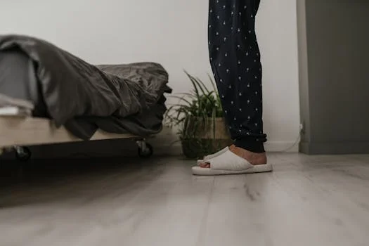 A person in pajamas and slippers stands on a wood floor next to a bed, suggesting when to see a podiatrist for heel pain.