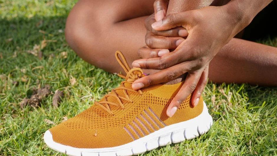 Athlete seated on grass holds the ankle above an orange running shoe.